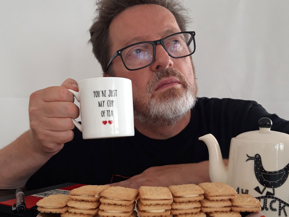 Poet Paul Cookson, raising a cup of tea, behind a wall of custard cream biscuits