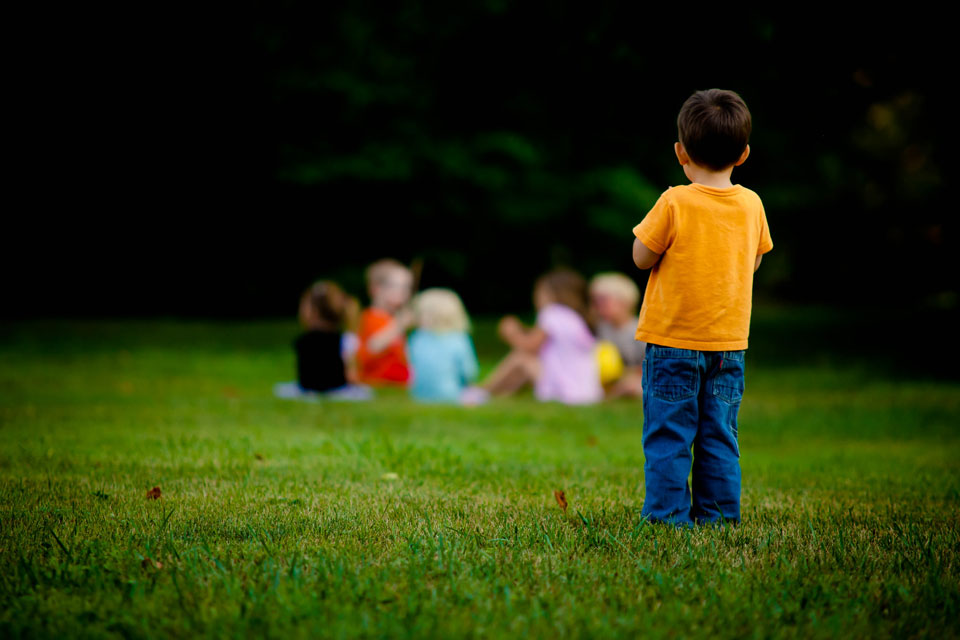 Young child standing isolated from a group playing on grass in front of him
