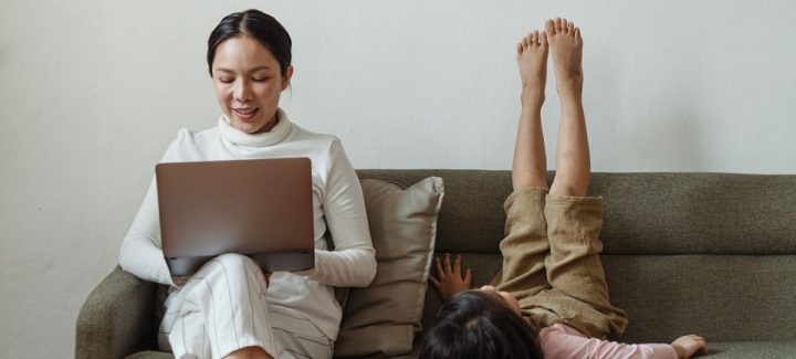 parenting_960 Mother sitting on a sofa looking at her laptop, child next to her is lying on sofa with feet in the air